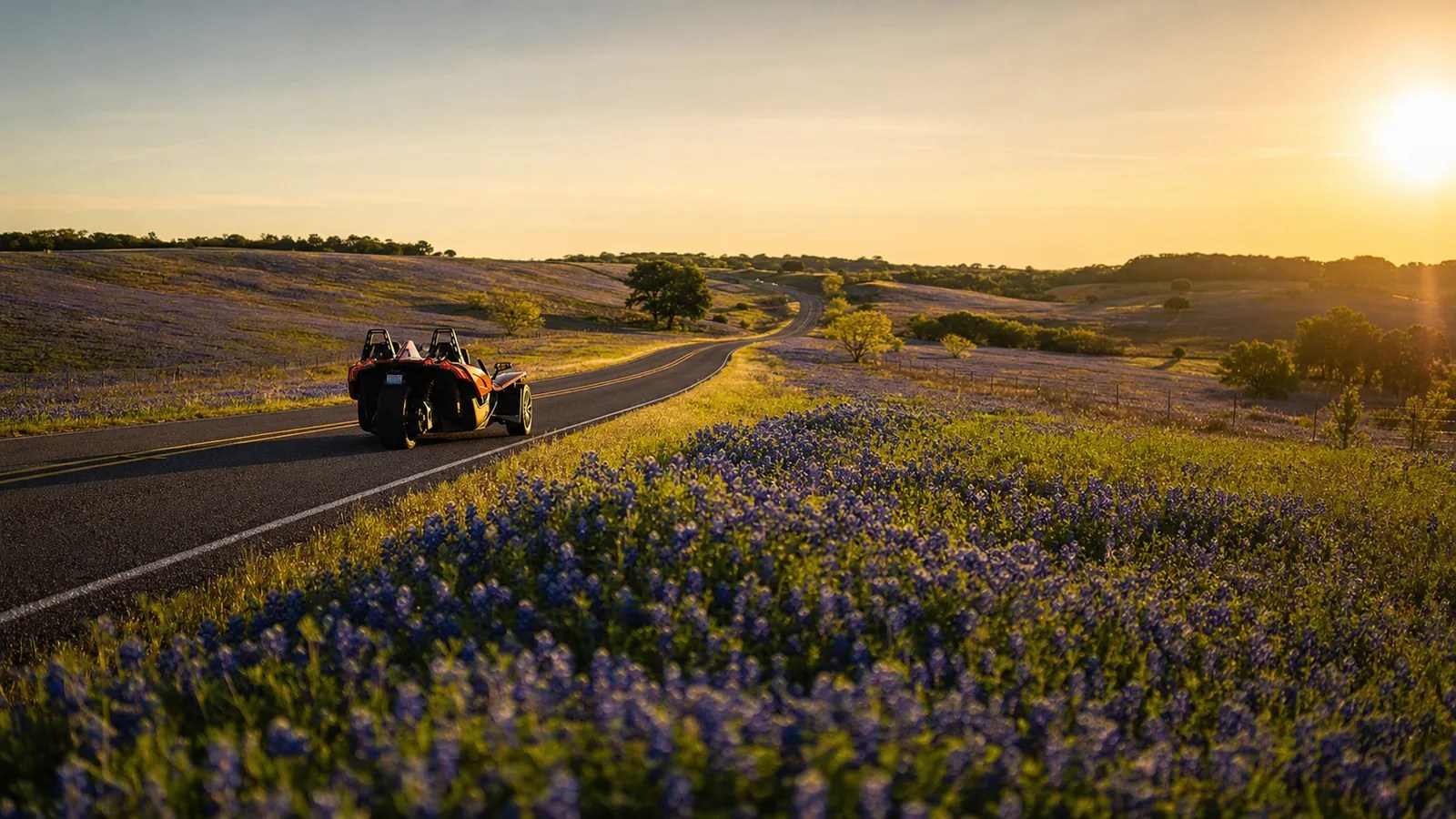 A winding Texas country road through bluebonnet fields with an orange Slingshot driving along it at golden hour. Hero image for the Ennis Slingshot article "How the Ennis Bluebonnet Trails Actually Work: 3 Loops, 40 Miles, One Simple System"