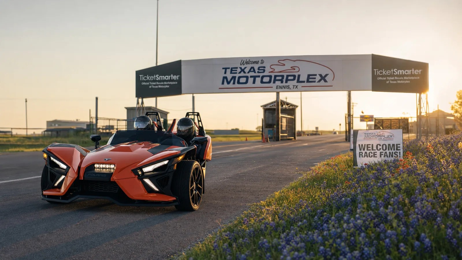 An orange Polaris Slingshot approaching the Texas Motorplex entrance gates at golden hour. Hero image for the Ennis Slingshot article "Texas Motorplex Parking, Traffic and Gate Times: What Nobody Tells You"