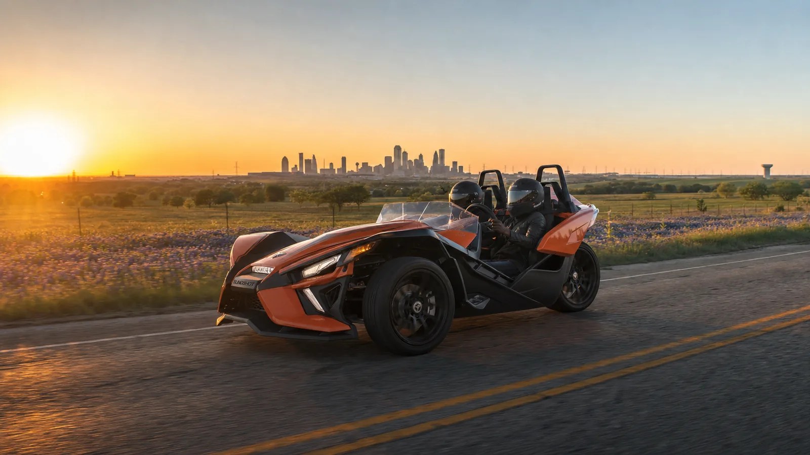 An orange Polaris Slingshot on a road with the Dallas skyline in the distance at golden hour. Hero image for the Ennis Slingshot article "Things to Do in Dallas This Weekend (Updated Weekly)"