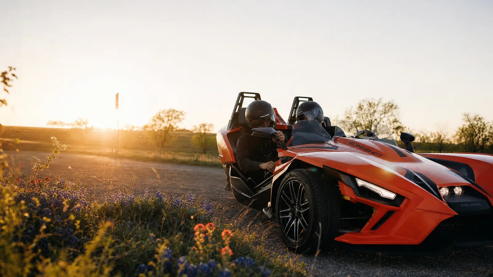 A helmeted driver adjusting the mirror of an orange Polaris Slingshot at golden hour. Hero image for the Ennis Slingshot article "10 Slingshot Safety Tips We Cover in Every Orientation"