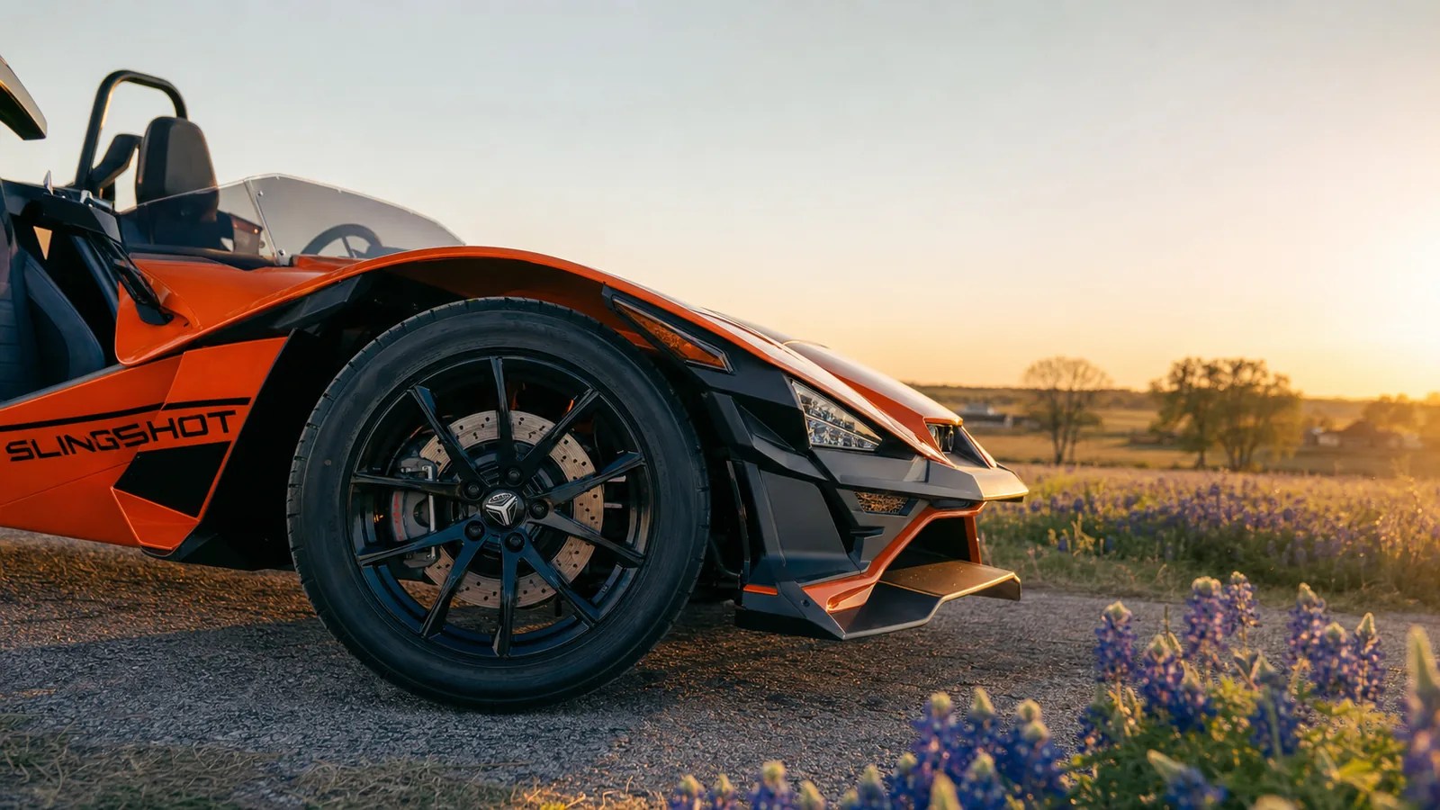 A close detail of the front wheel and brake of an orange Polaris Slingshot at golden hour. Hero image for the Ennis Slingshot article "How Polaris Slingshot Brakes Feel (and Why to Leave More Room Than You Would in a Car)"