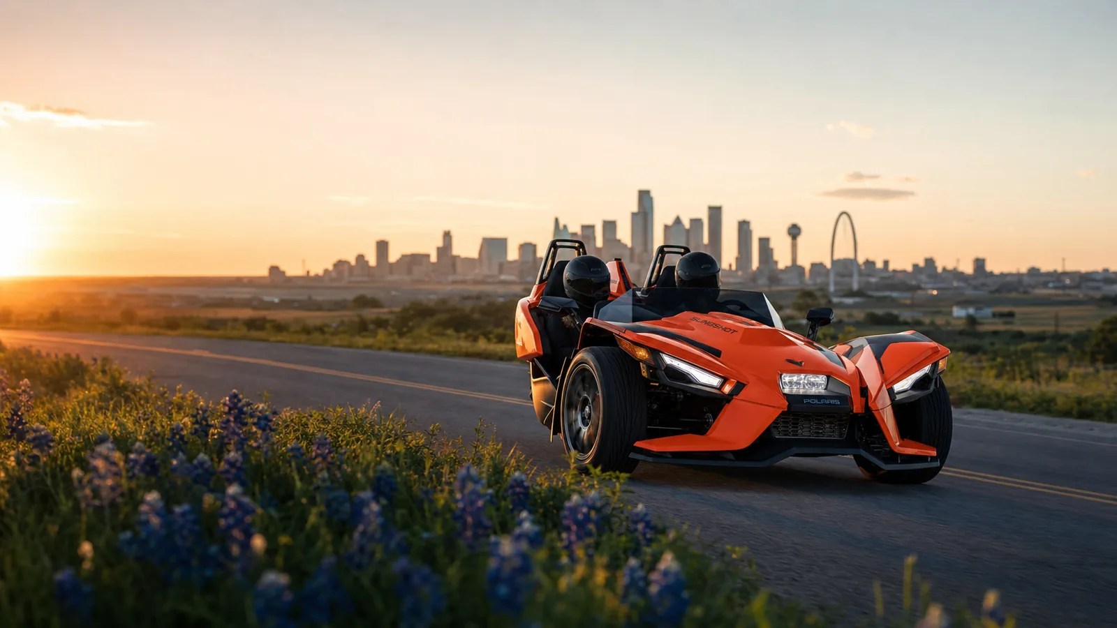 An orange Polaris Slingshot on a road near Dallas landmarks at golden hour. Hero image for the Ennis Slingshot article "The Dallas Bucket List: Things Every Couple Should Do"