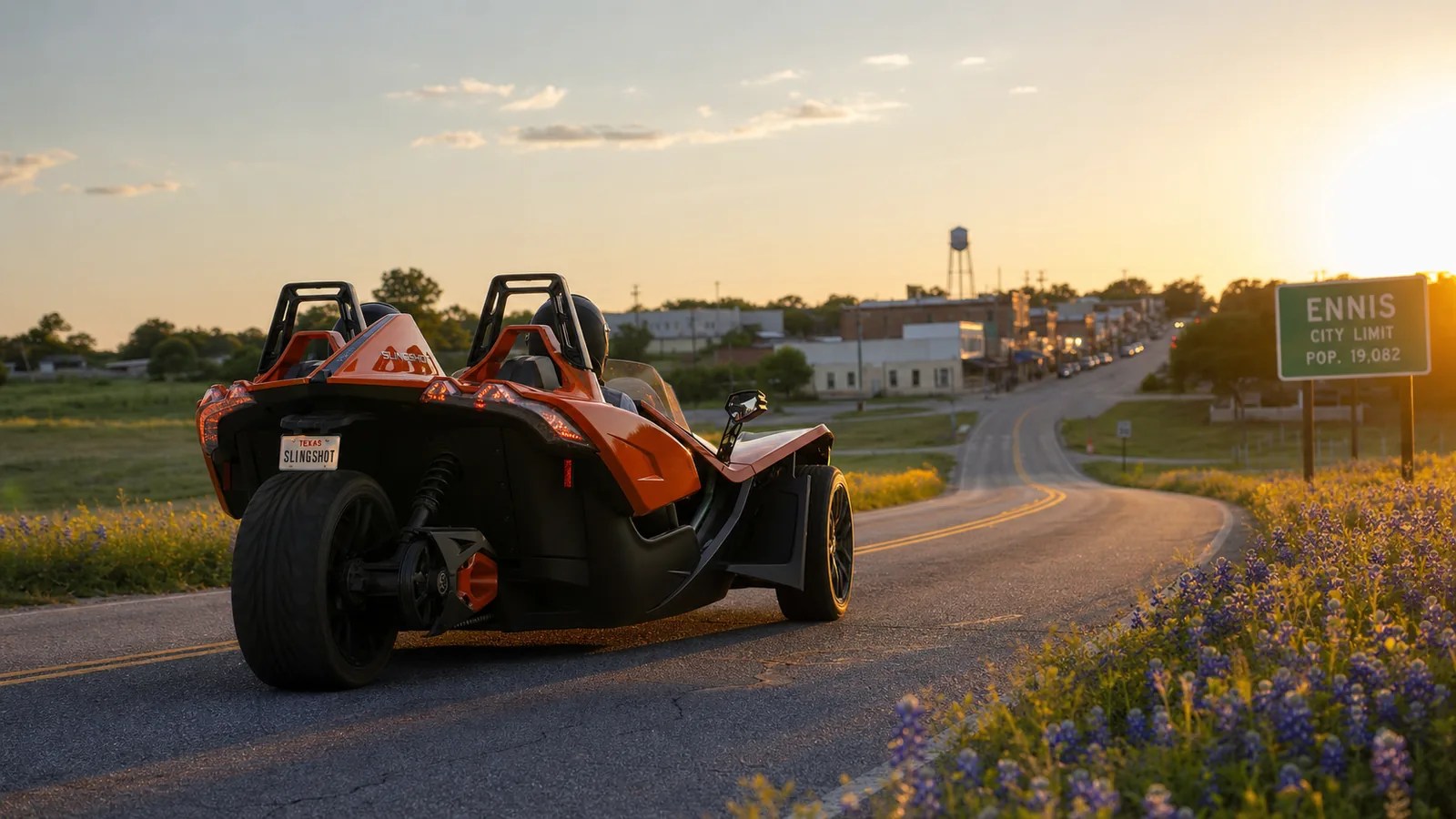 An orange Polaris Slingshot driving toward a small Texas town at golden hour. Hero image for the Ennis Slingshot article "Small Towns Near Dallas Worth a Day Trip (Ranked by Drive Time)"