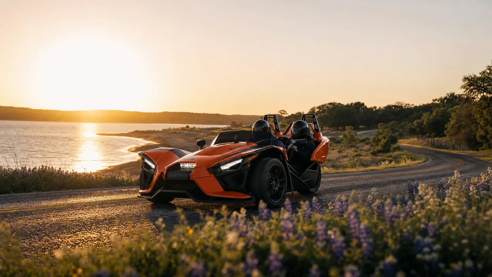 An orange Polaris Slingshot on a scenic lakeside drive at golden hour. Hero image for the Ennis Slingshot article "Scenic Drives Near Dallas: Roads Worth the Gas (Year-Round)"